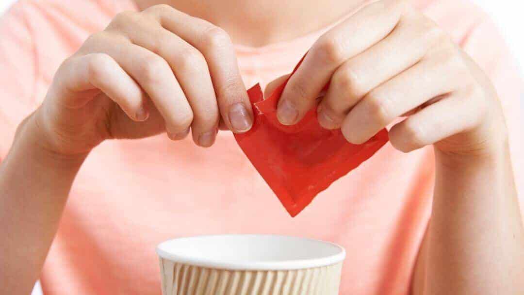 Hands tearing a red paper heart above a cup, symbolising the potential health dangers of artificial sweeteners.
