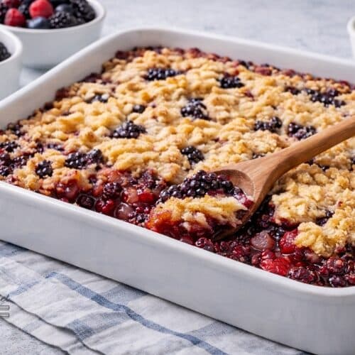 Low-Carb Raspberry & Blackberry Cobbler in a white baking dish on a kitchen table with a wooden spoon serving the berry crumble and fresh berries nearby