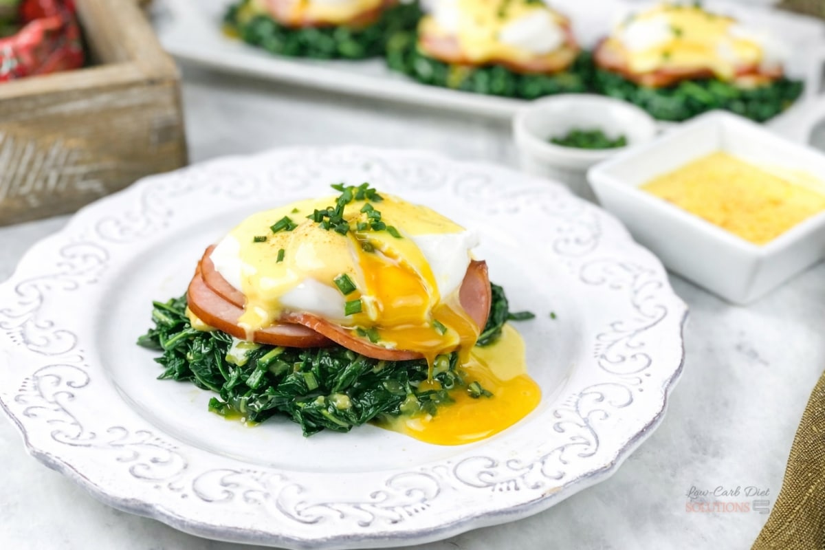Keto Eggs Benedict with Spinach and Hollandaise 1 Low-carb keto eggs benedict served on sautéed spinach with Canadian bacon, poached egg, and creamy hollandaise sauce on a white plate